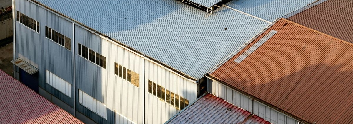 An aerial view of a large industrial warehouse facility with corrugated metal roofing panels in shades of grey and brown.