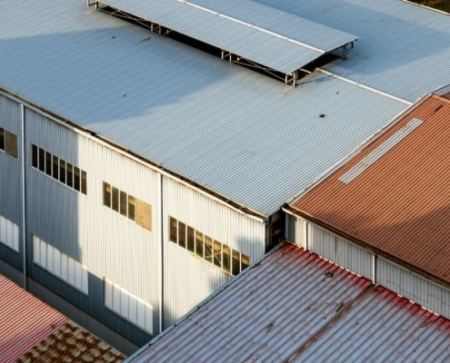 An aerial view of a large industrial warehouse facility with corrugated metal roofing panels in shades of grey and brown.