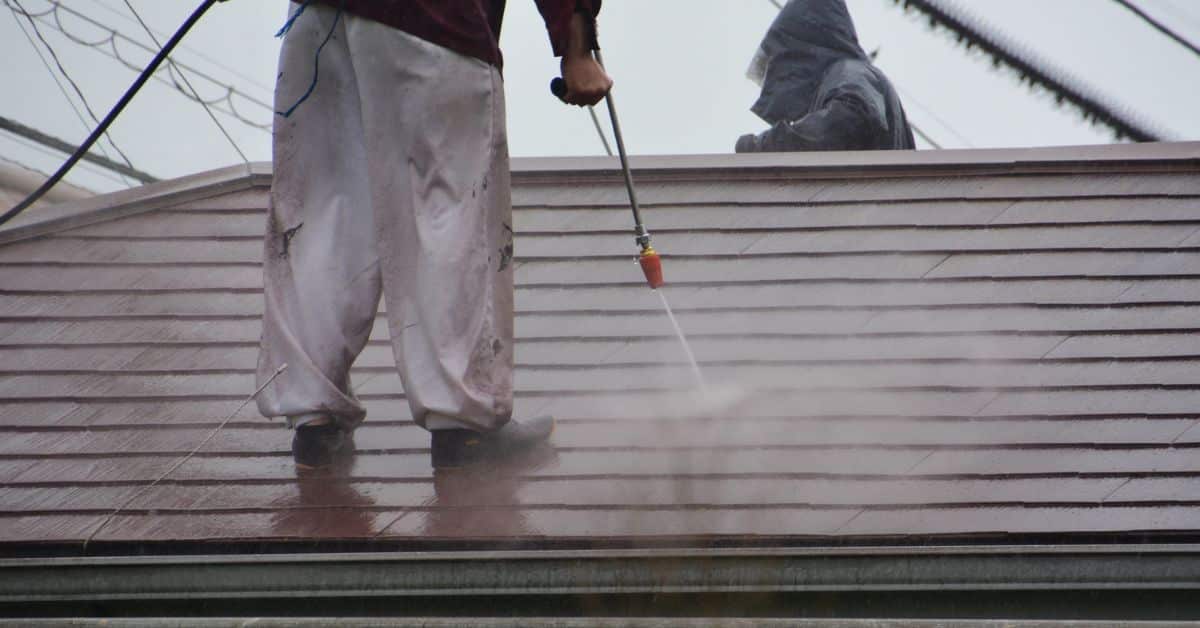 A technician cleaning a brown tiled roof using a high-pressure water sprayer wand.