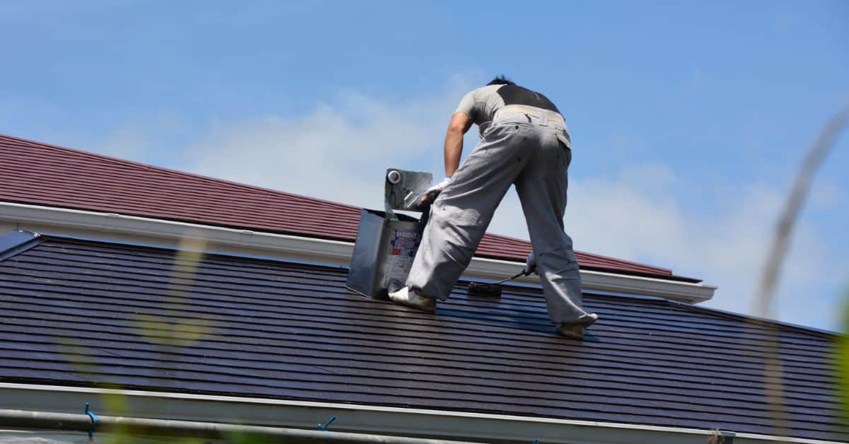 A worker in protective gear applying a protective roof coating with a roller to a dark shingled roof.