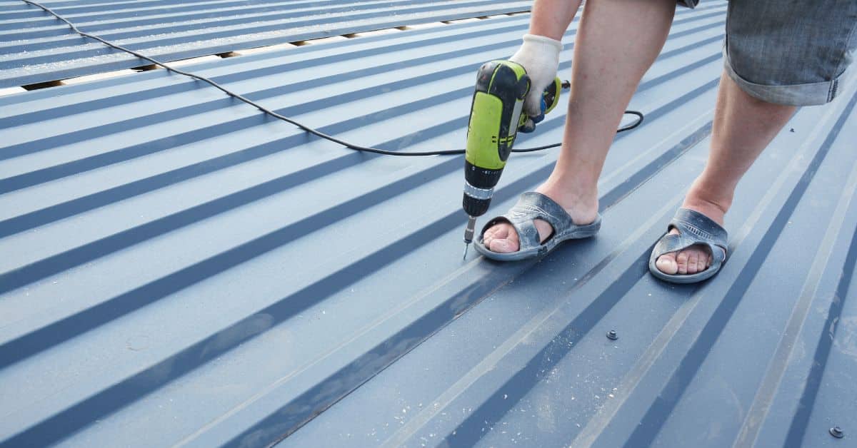 Close-up of a worker using a cordless drill to secure specialized roofing screws into the ribs of a blue corrugated metal panel.