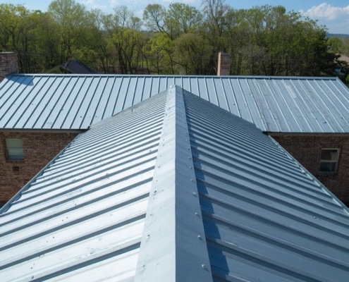 A top-down, aerial photograph taken from the perspective of a roofer. The image focuses on a newly installed light blue, vertically-ridged metal roof structure, highlighting the clean alignment and fastening patterns essential for a successful metal roof installation.