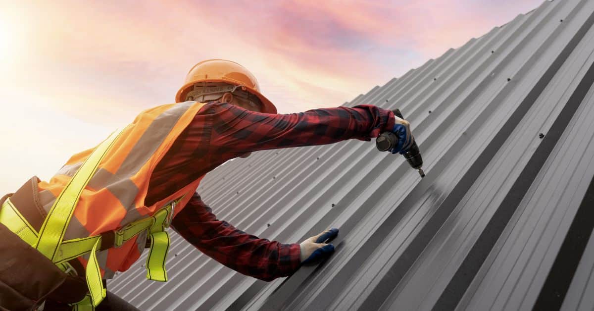 An HVAC or roofing professional in high-visibility gear and a hard hat using a power tool to install a grey standing seam metal roof at sunset.