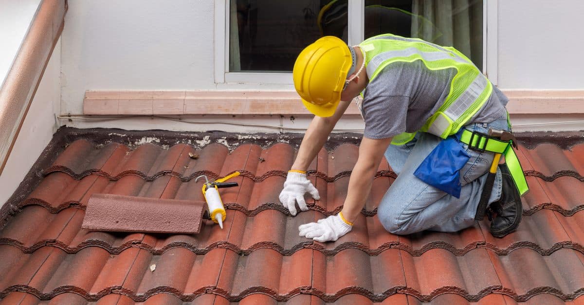 A roofing technician in a safety vest and yellow hard hat applying sealant to red barrel roof tiles near a wall flashing point to prevent leaks.