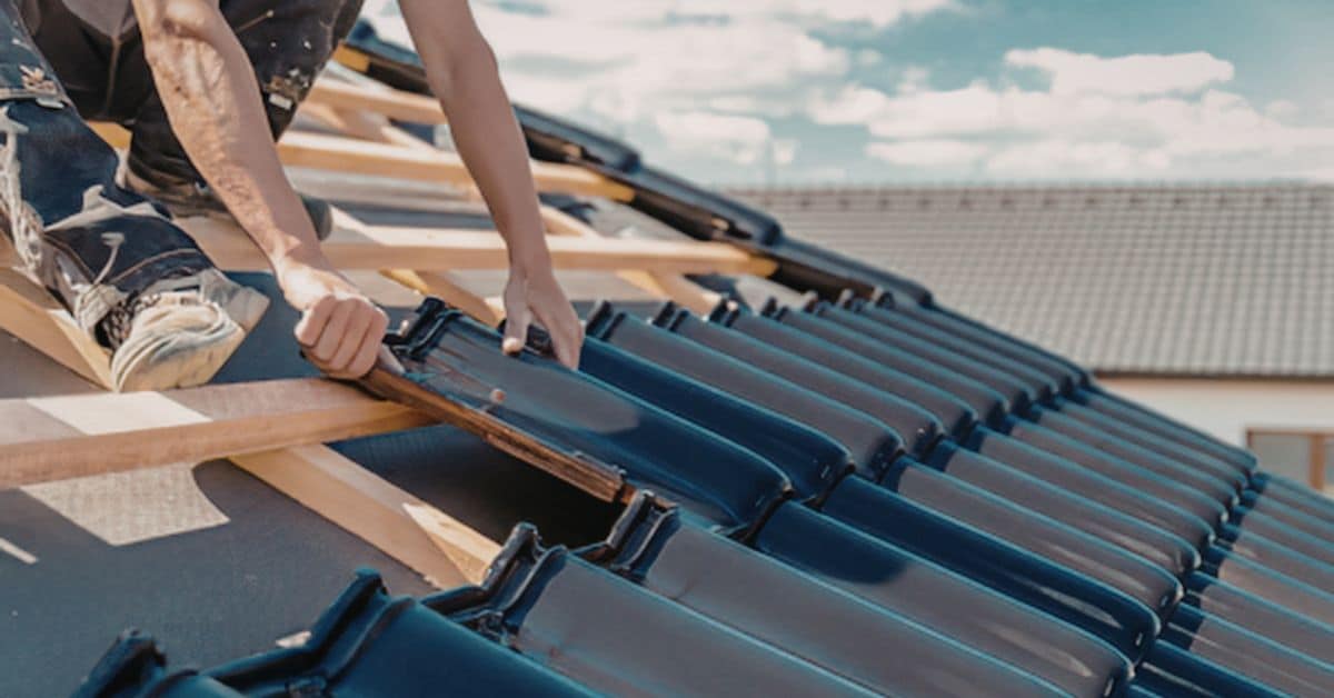 Professional roofer's hands carefully sliding a black interlocking roof tile into place on a timber batten framework during a full roof replacement.