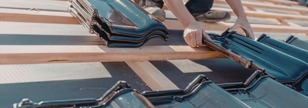 A close-up of a roofer's hands precisely aligning new black interlocking roof tiles over a wooden batten system and waterproof underlayment.