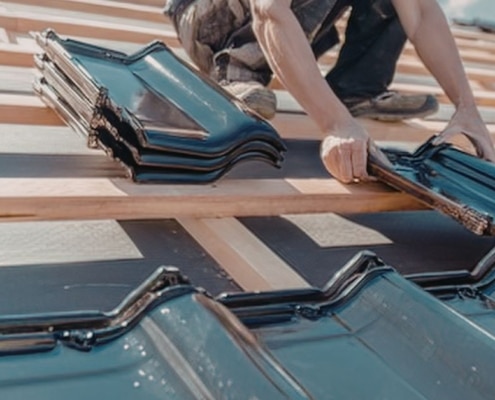 A close-up of a roofer's hands precisely aligning new black interlocking roof tiles over a wooden batten system and waterproof underlayment.